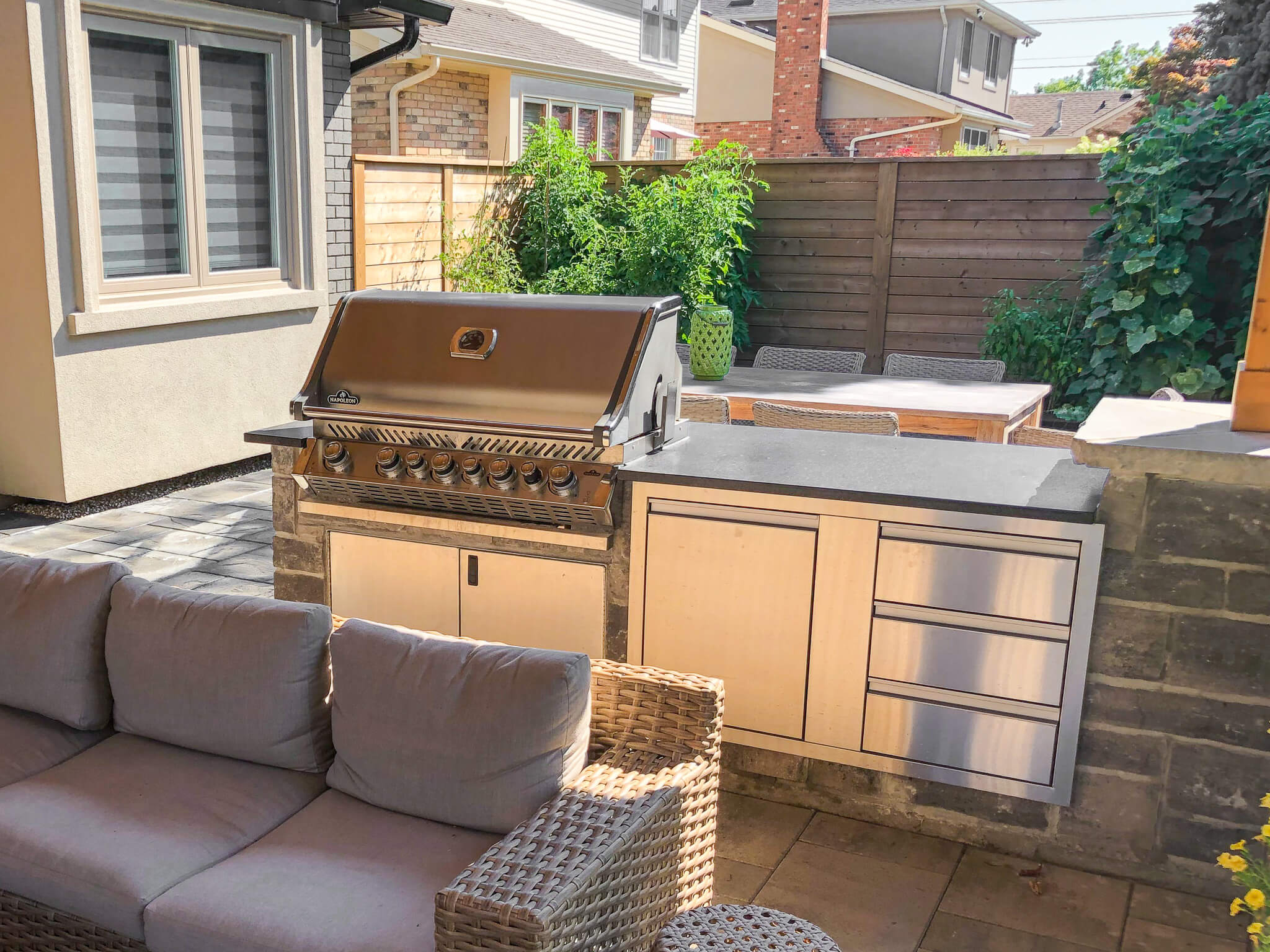 Outdoor kitchen design with stainless Napoleon grill, stone cabinetry, and nearby lounge seating under natural sunlight.