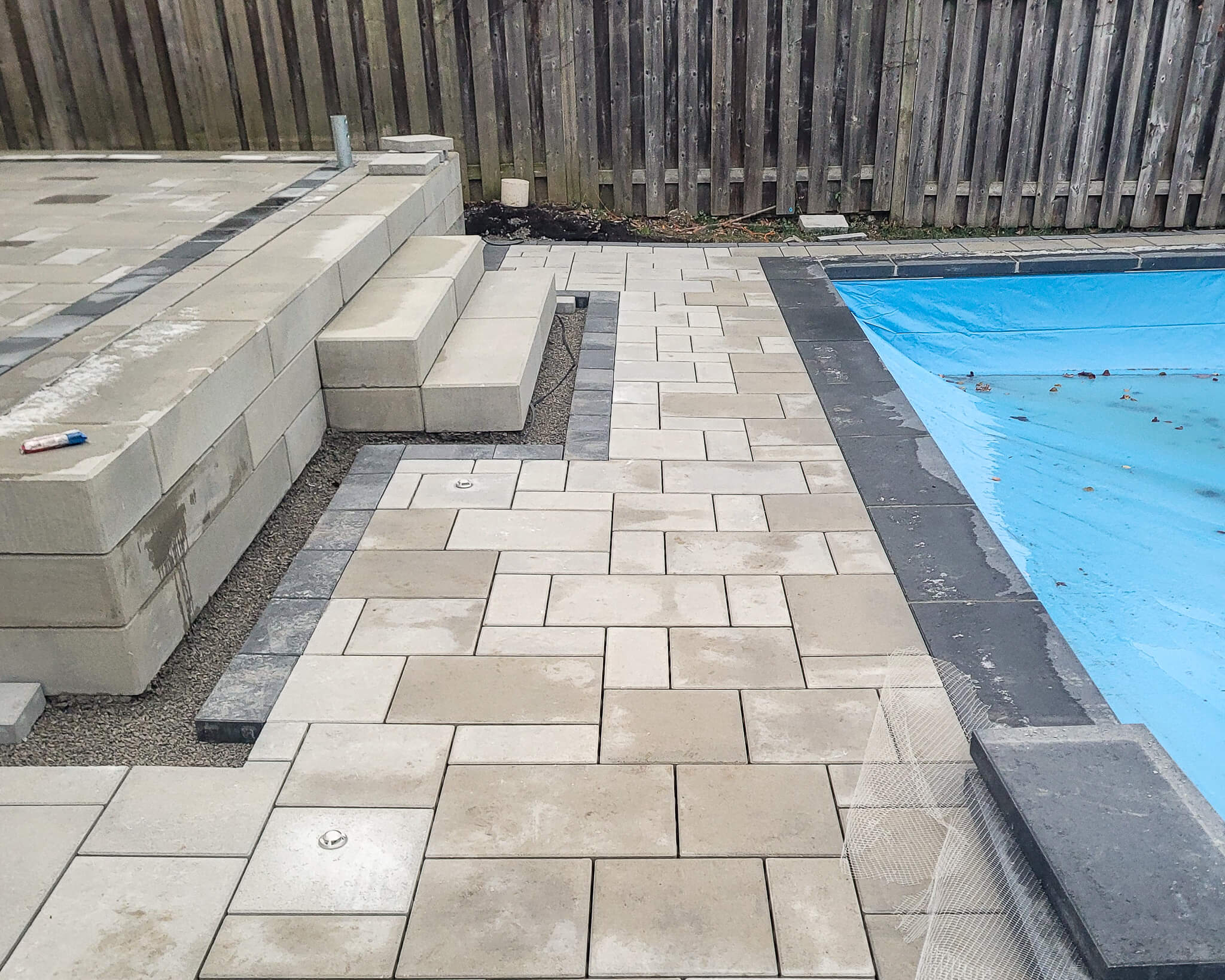 Poolside patio pavers installed with integrated steps, retaining wall blocks, and dark border accents beside a covered pool