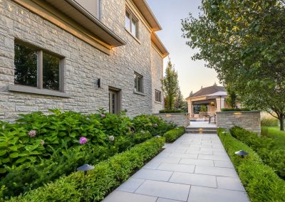 A structured stone walkway framed by layered planting and subtle lighting, guiding movement through a refined residential landscape.