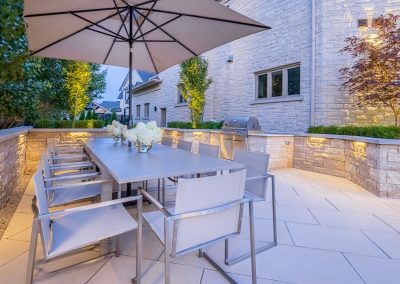 An open-air dining terrace designed for entertaining, featuring integrated lighting, built-in grilling, and clean stone detailing that extends outdoor living into the evening.