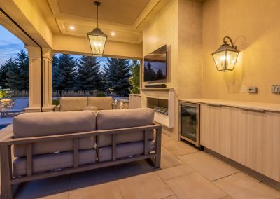 A covered outdoor lounge features integrated seating, a modern fireplace, built-in cabinetry, and warm architectural lighting, creating a comfortable space overlooking the pool and surrounding landscape.