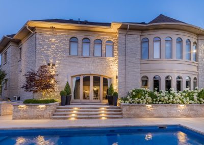 A refined backyard poolscape features a stone home facade, tiered entry steps, integrated lighting, and layered planting beds that frame the outdoor living space.