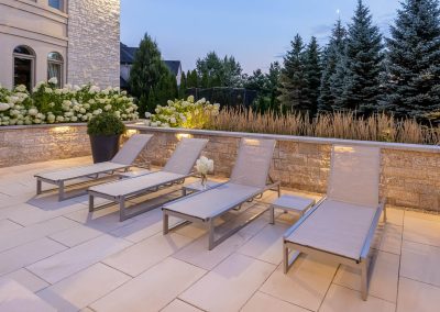 A clean-lined poolside lounge area features stone retaining walls, integrated lighting, and layered planting that creates a calm, resort-inspired backyard setting.