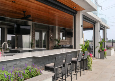 Covered outdoor kitchen featuring a long stone bar, modern cabinetry, wood ceiling, bar seating, and surrounding planters.