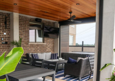 A covered backyard lounge area with wicker seating, accent pillows, a mounted outdoor TV, brick feature wall, and wood-paneled ceiling, offering a comfortable space beside the outdoor kitchen.