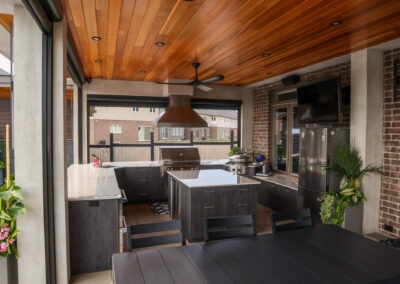 A detailed view of a premium outdoor kitchen showcasing custom dark cabinetry, stainless steel grill and appliances, a large central island, ceiling fan, and natural wood ceiling within a covered entertainment pavilion.