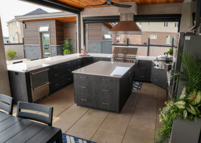 A spacious luxury outdoor kitchen featuring dark modern cabinetry, sleek waterfall-edge countertops, stainless steel appliances, a large island, and tile flooring, designed as part of a covered backyard entertaining space.