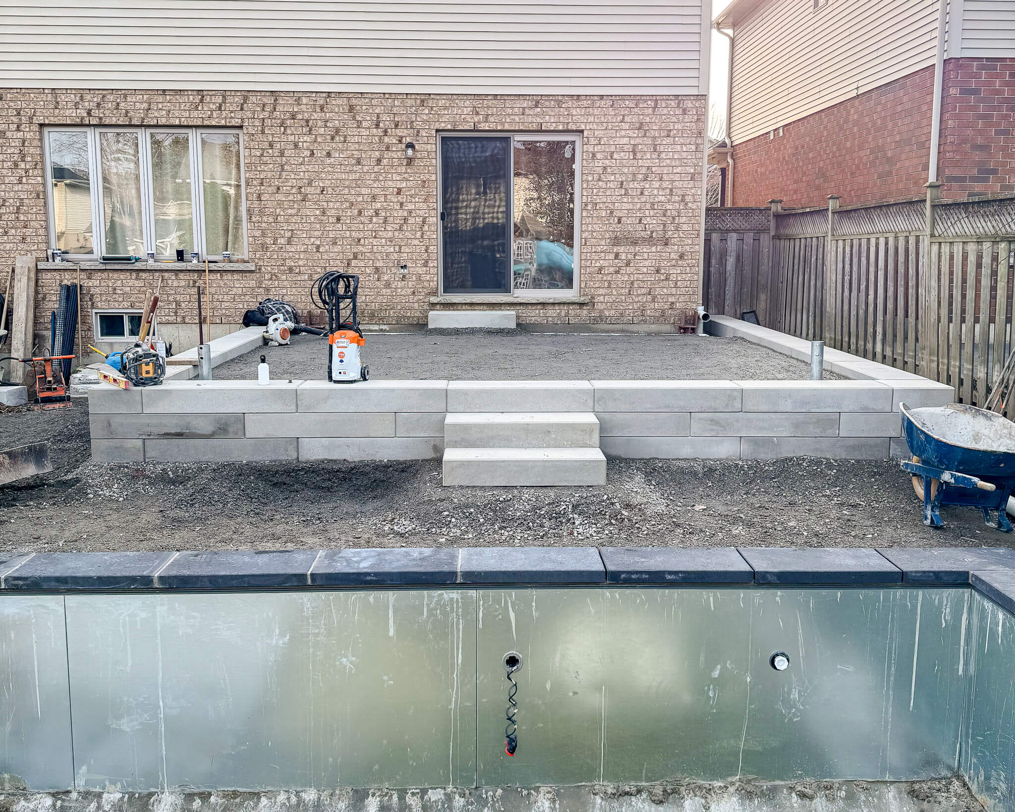 Raised patio under construction with concrete steps, retaining wall blocks, and compacted base material above a new pool