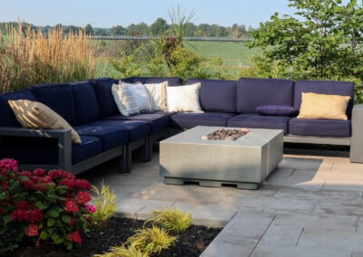 Sectional outdoor seating surrounding a Sanctuary concrete fire table, framed by shrubs, grasses, and countryside views.