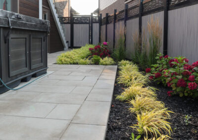 Pathway paved with beige slabs beside a hot tub, lined with ornamental grasses, hydrangeas, and modern fencing.