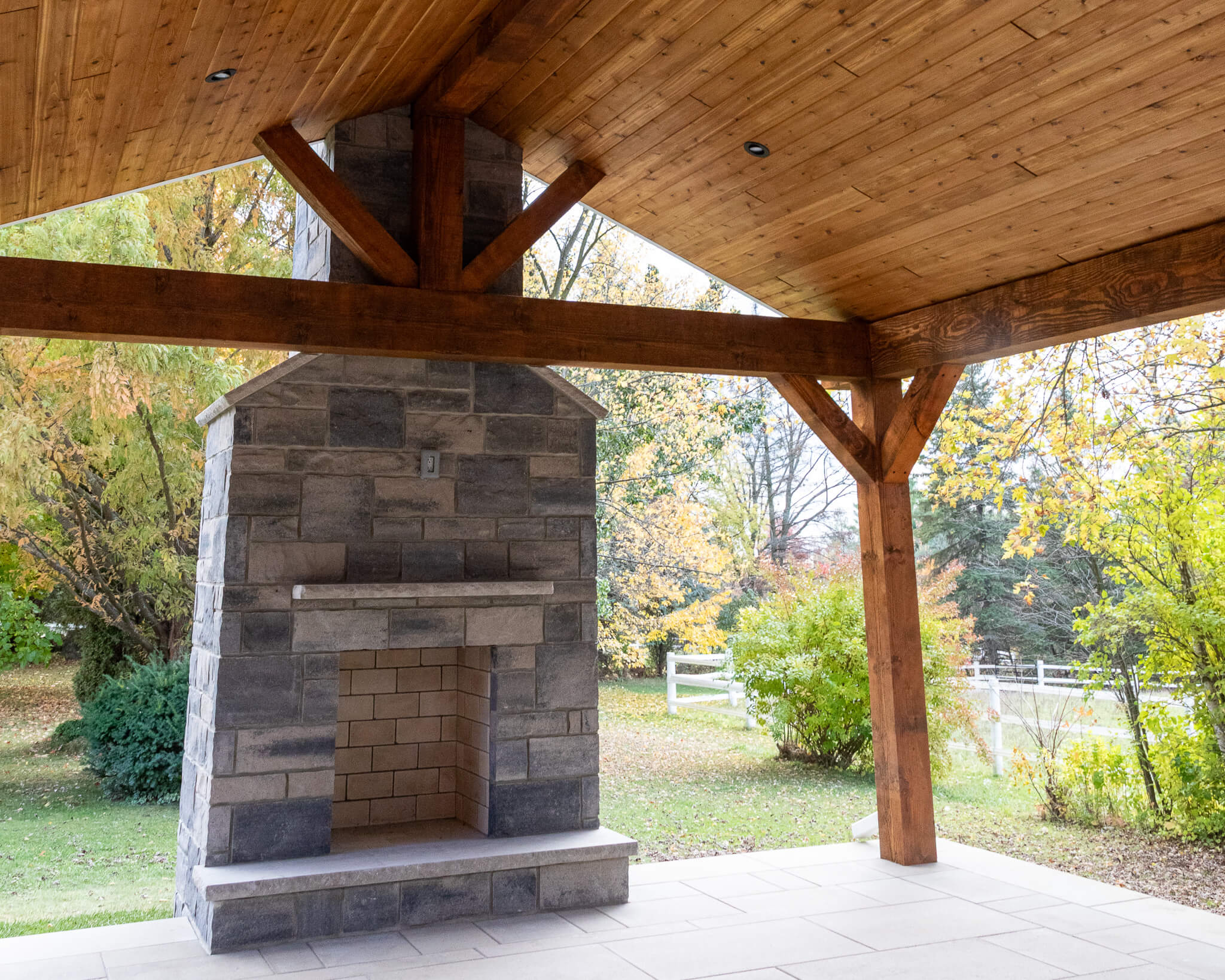 Wood-framed covered pavilion with a stone wood-burning outdoor fireplace, finished with natural stone masonry and installed on a large slab patio in a residential backyard.