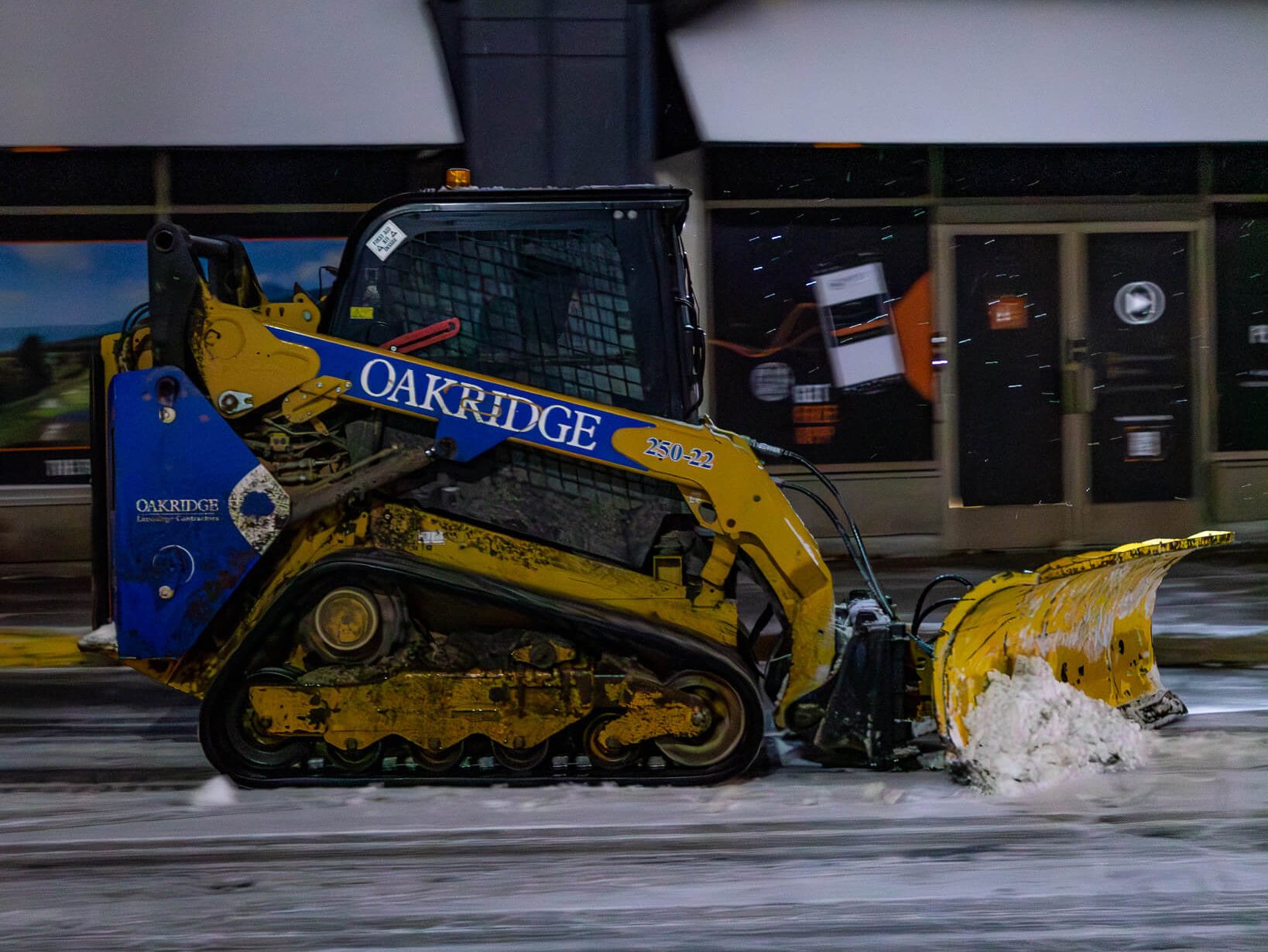 Oakridge Snow Clearing Operations in Active Winter Conditions Oakridge skid steer plowing snow in a commercial parking area during active winter weather conditions.
