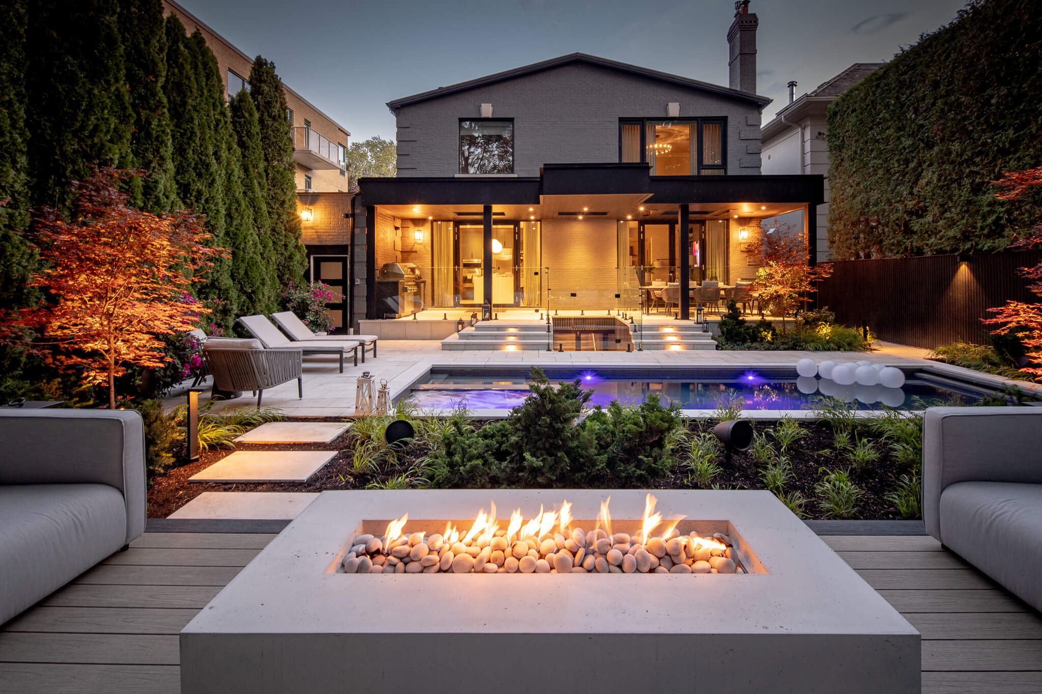 Modern backyard featuring a rectangular gas fire feature in the foreground, integrated pool, landscaped seating areas, and a covered patio with outdoor living spaces illuminated at dusk.