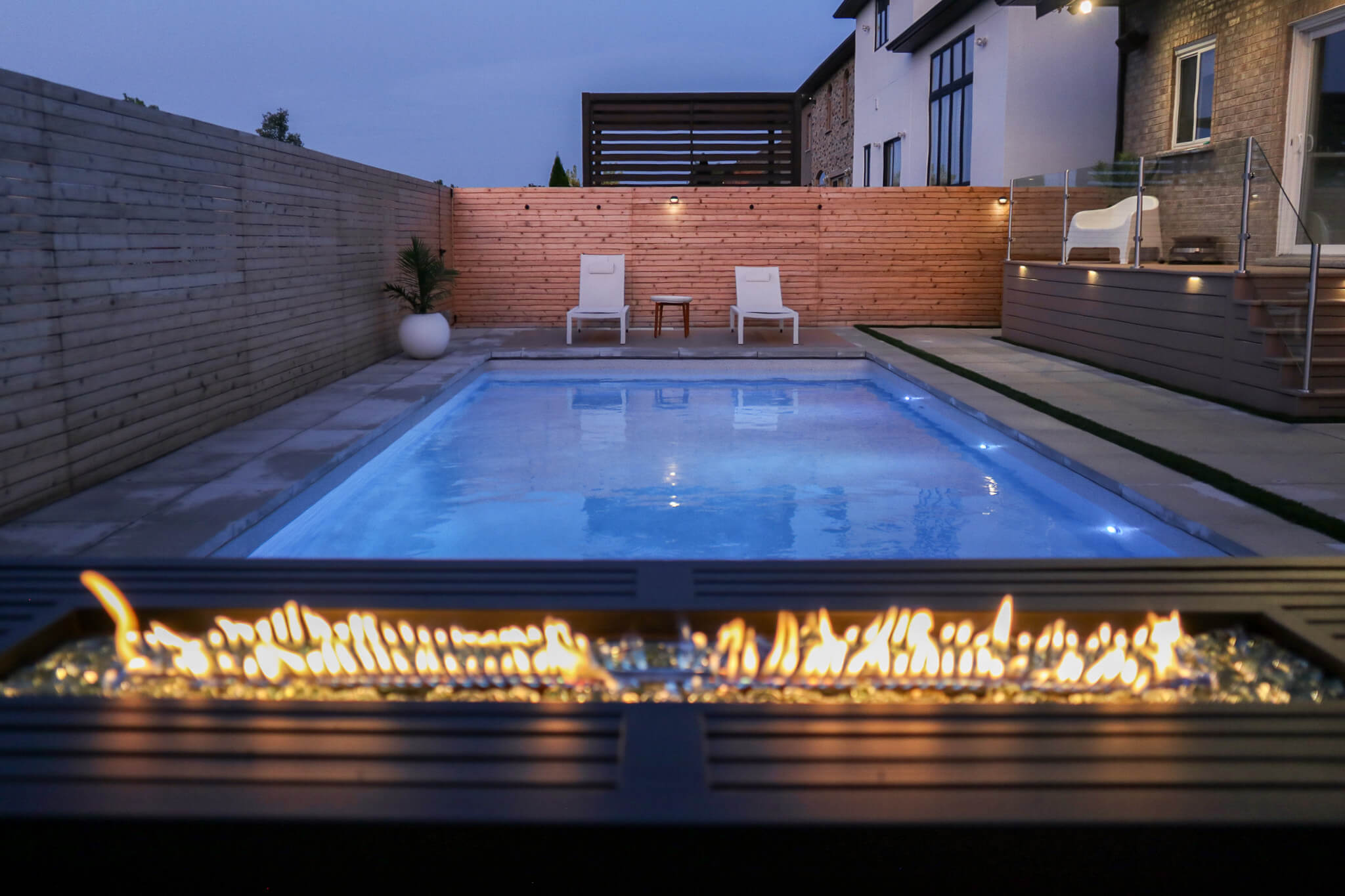 Linear gas fire table in the foreground overlooking a modern swimming pool and patio with integrated evening lighting and privacy fencing