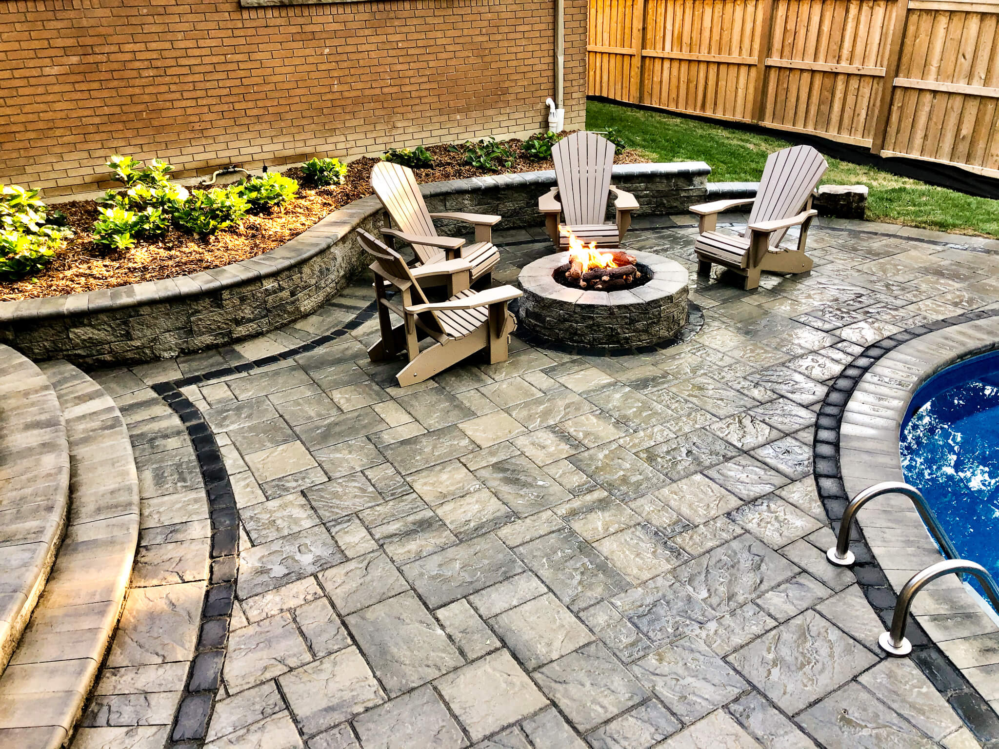 Stone fire pit surrounded by Adirondack chairs on a poolside patio with curved retaining wall and interlock paving